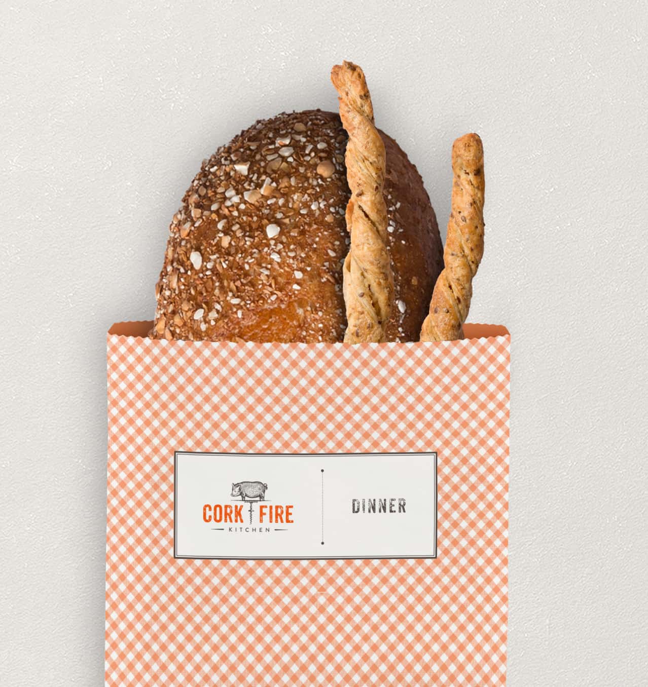A paper bag with a checkered orange pattern hanging on the wall containing a round loaf of bread and two breadsticks, bearing the 'CORK & FIRE' logo.