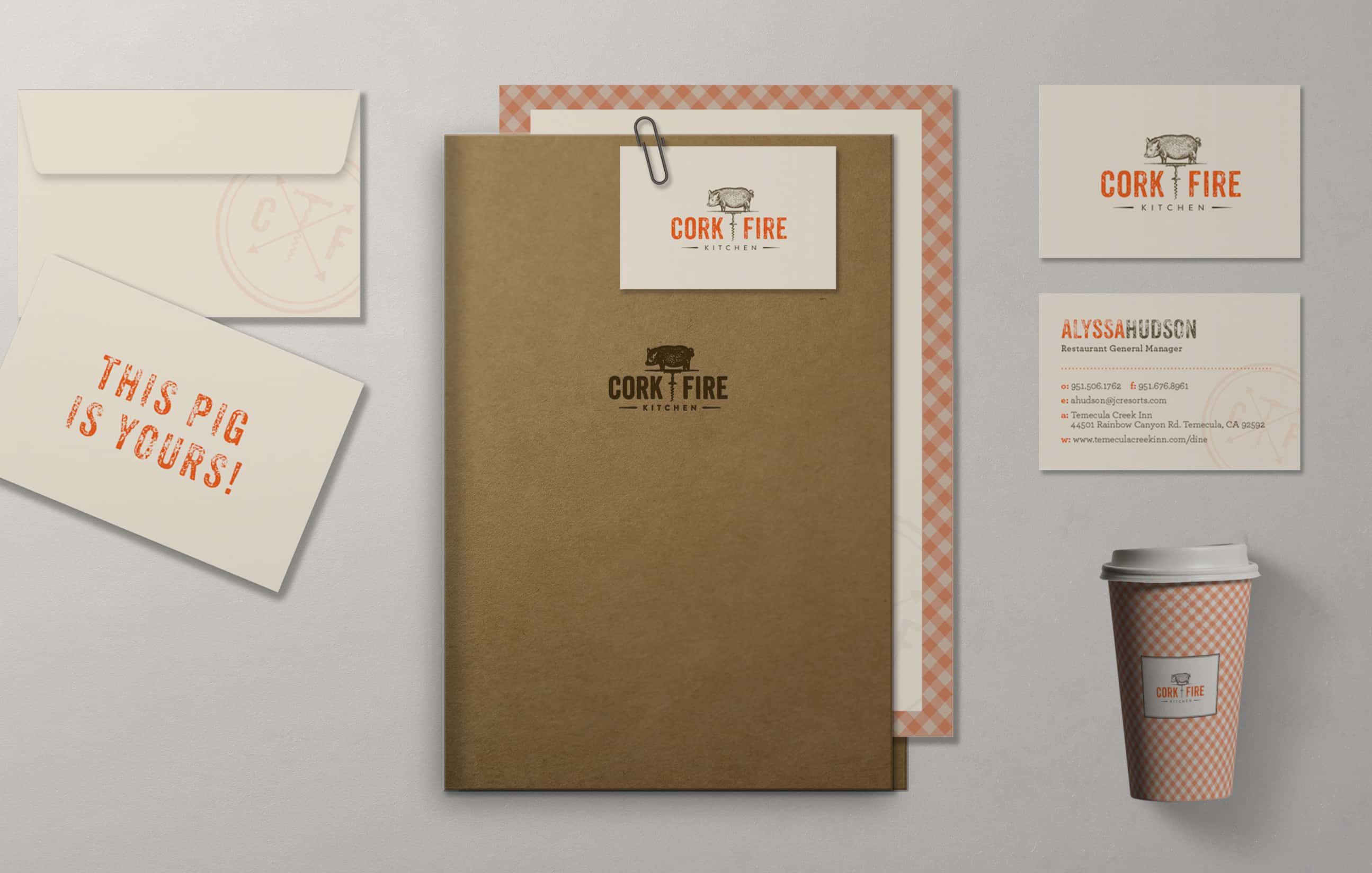 Branding materials for 'CORK & FIRE' displayed on a wall, including a kraft paper menu, a business card, a coffee cup with a checkered pattern, and a promotional card stating 'This pig is yours!'