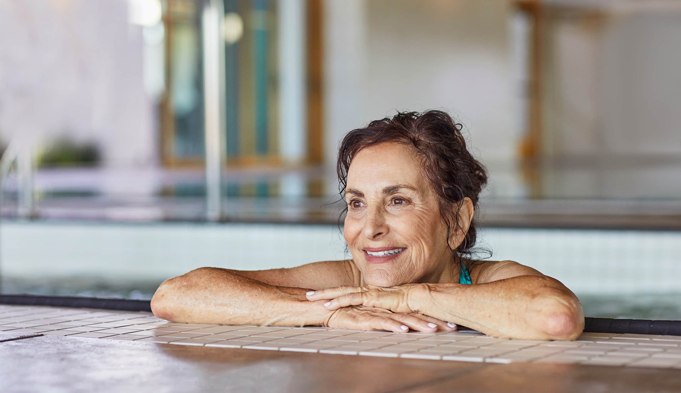 A senior woman with a content smile relaxing at the edge of an indoor swimming pool, suggesting an active and enjoyable retirement lifestyle.