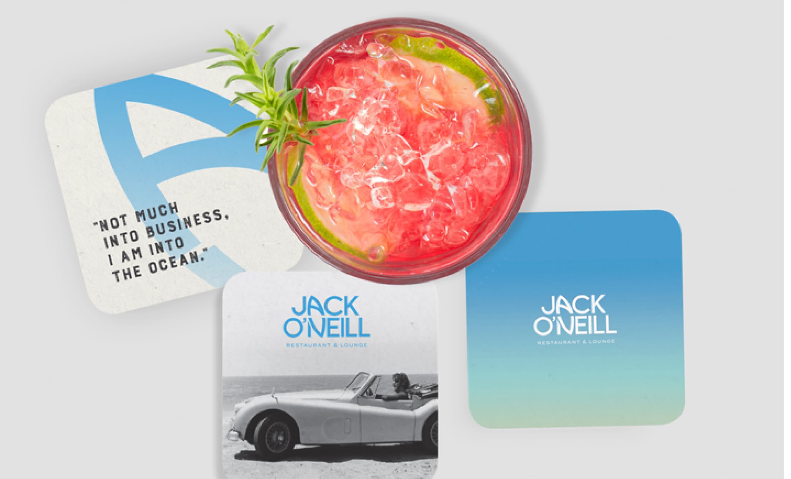 A beverage on a coaster with a quote saying 'NOT MUCH INTO BUSINESS, I AM INTO THE OCEAN.' beside two coasters with branding for Jack O'Neill Restaurant & Lounge and a vintage image of a convertible car on the beach.