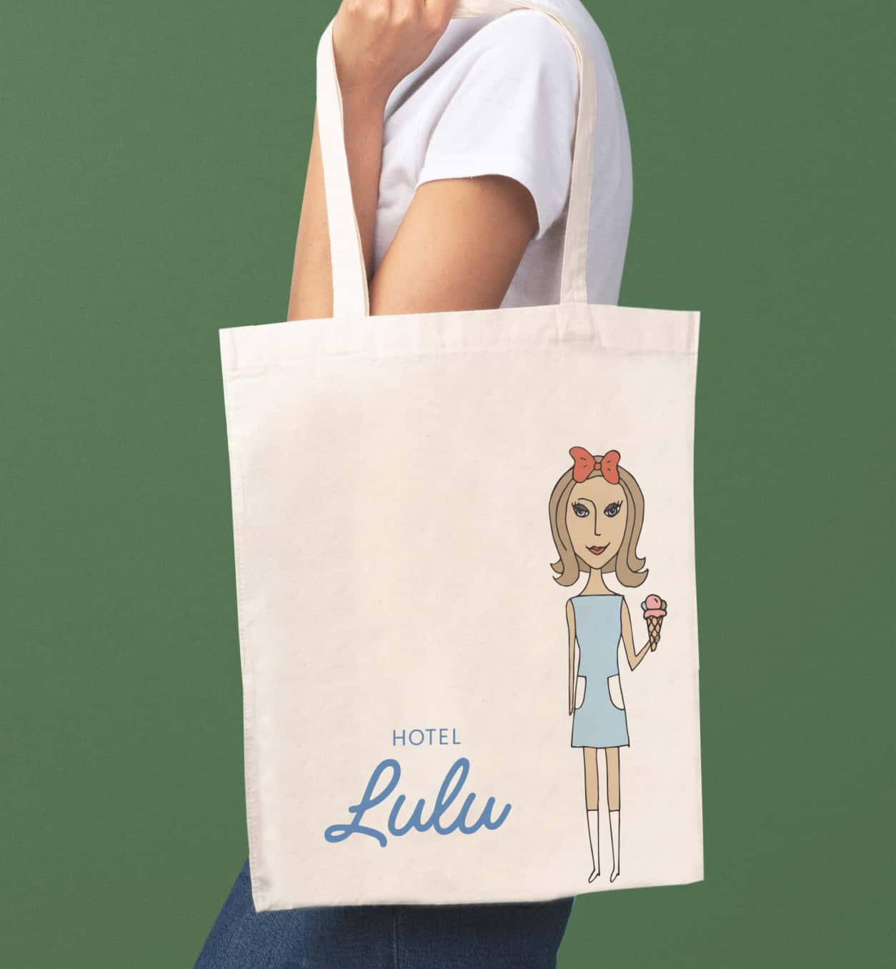 A person carrying a canvas tote bag with 'Hotel Lulu' branding, featuring a drawn female character with a hotel bellhop cart and luggage, indicative of hotel guest services.