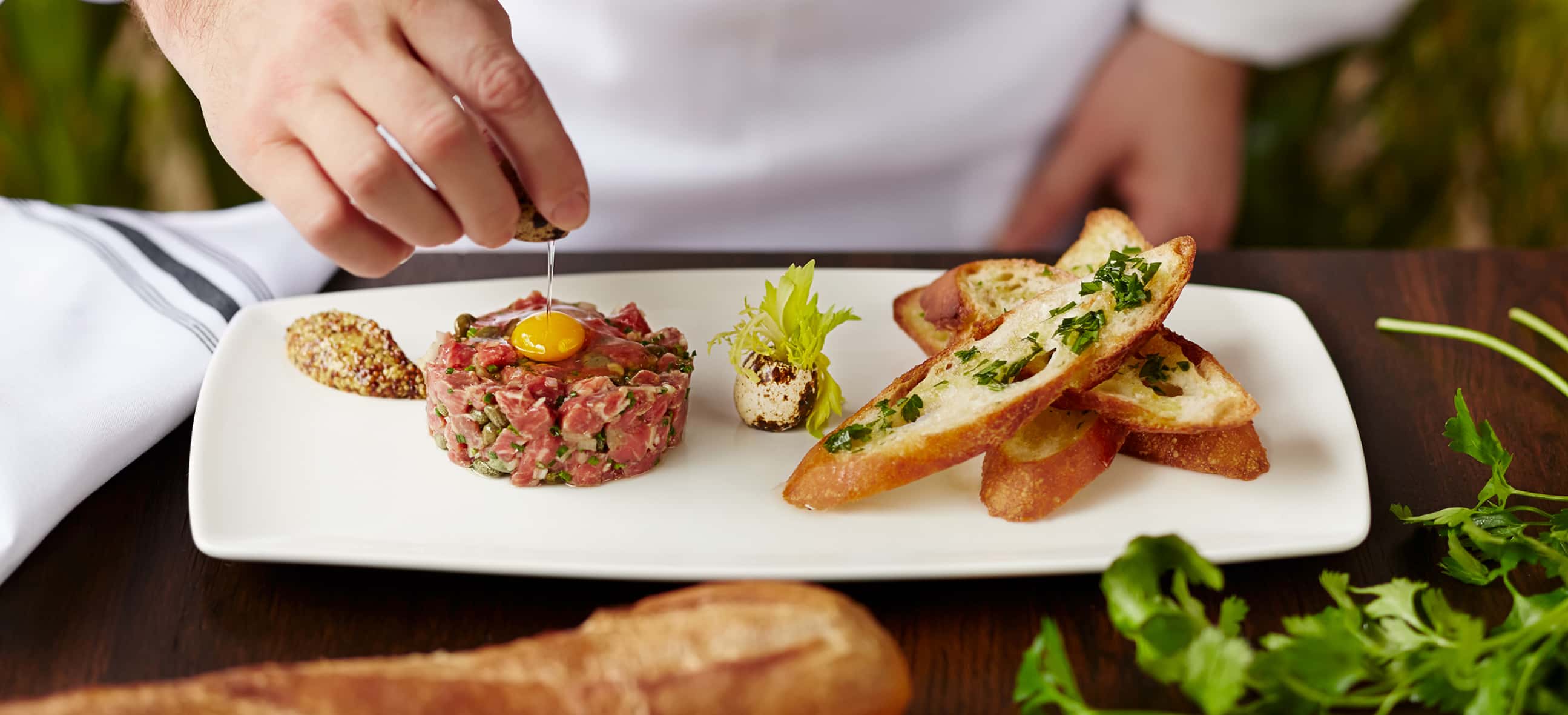 A chef in a white jacket is garnishing a dish of steak tartare with an egg yolk on top, accompanied by toasted bread and a side of seasoning.