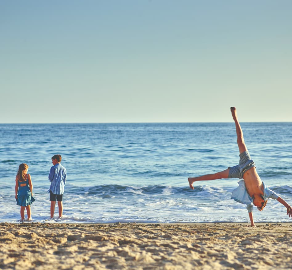 Two kids play in the water and one person does a handstand on the beach at Surf & Sand Resort